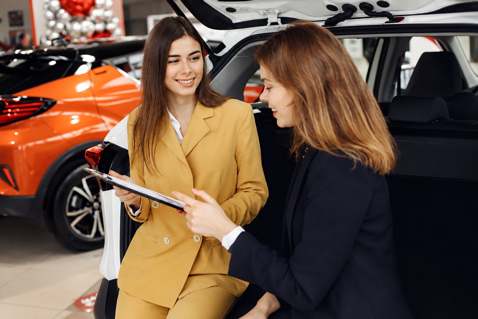 Twee zakelijke vrouwen bespreken documenten bij een auto in een showroom.